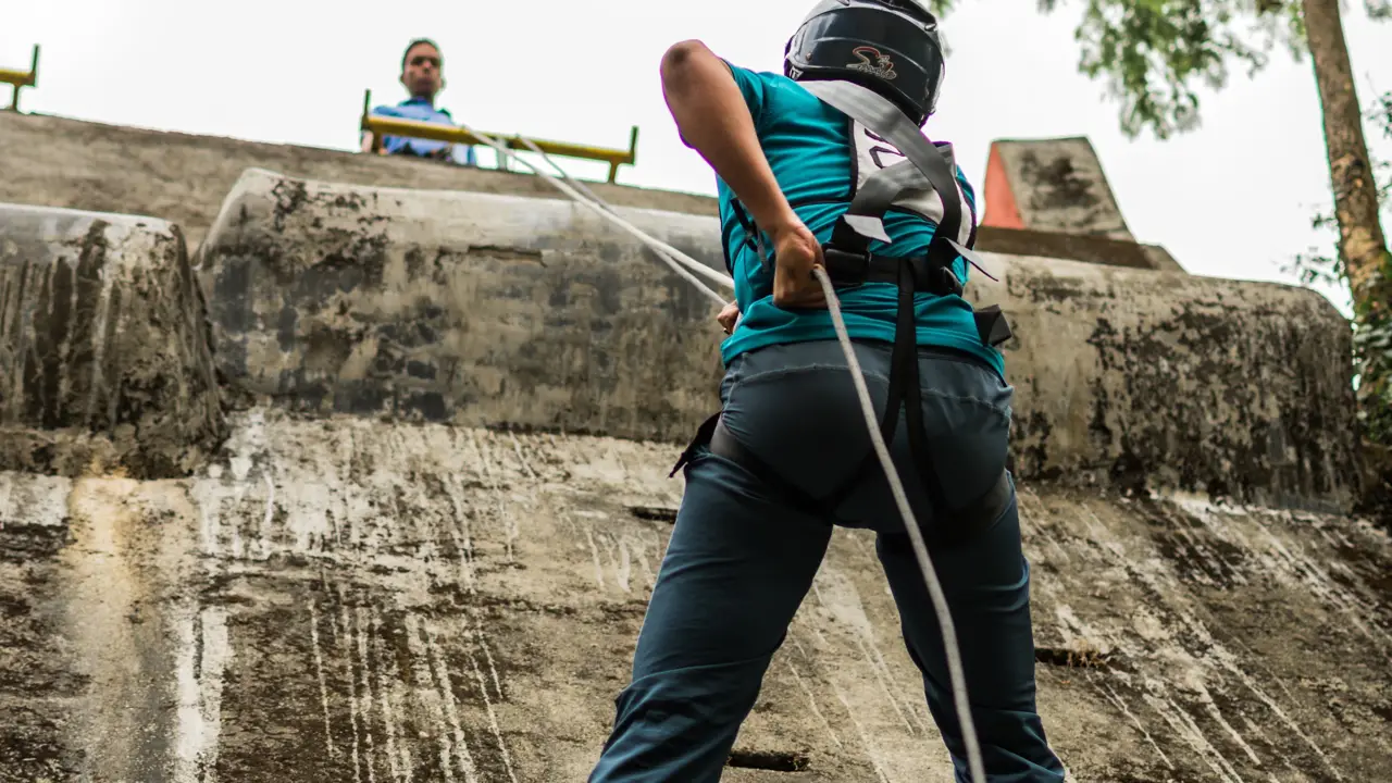 A person in a harness and helmet rappelling down a steep wall with a partner watching from the top