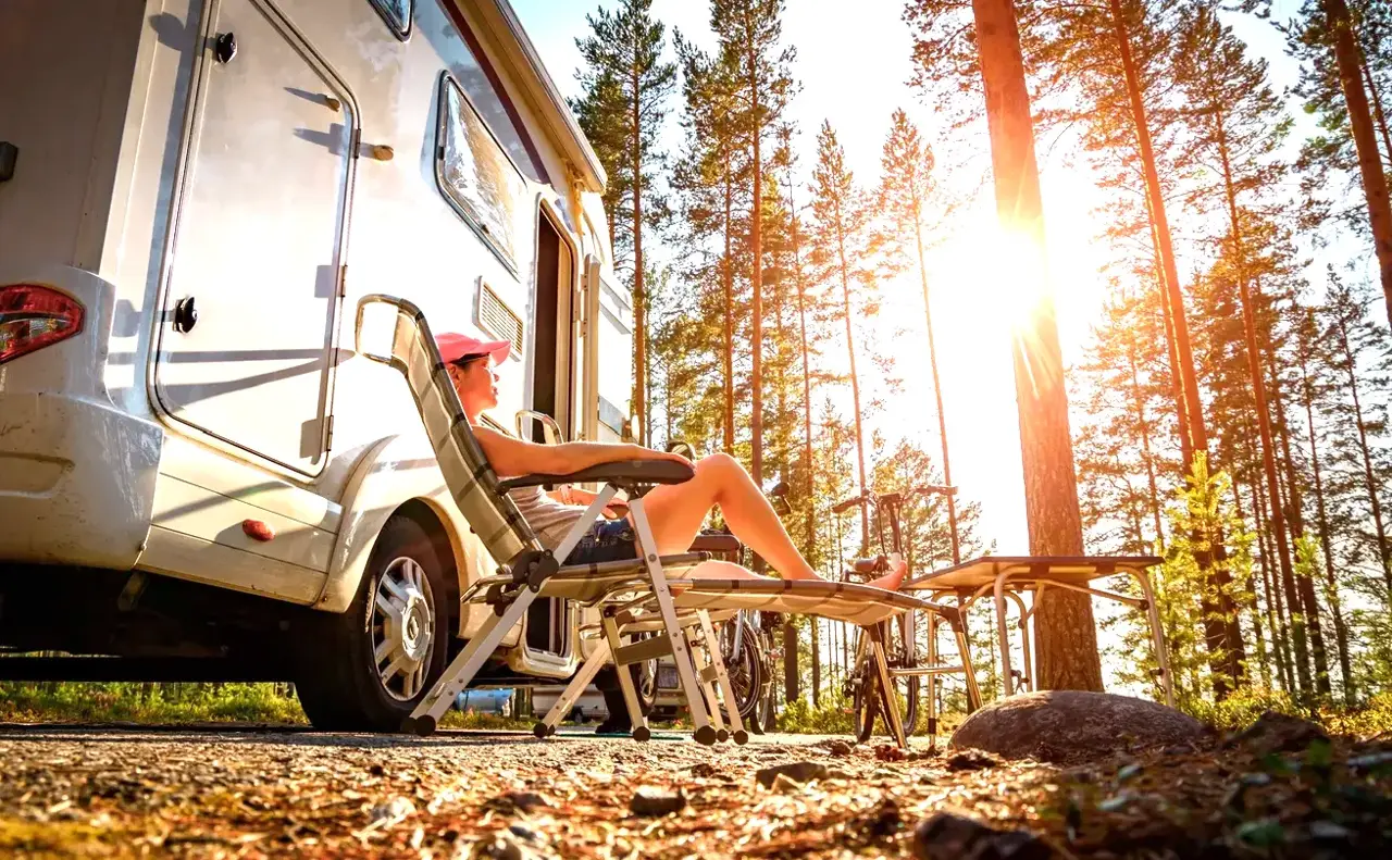 A camper relaxing in a lounge chair beside an RV among tall pines at sunrise